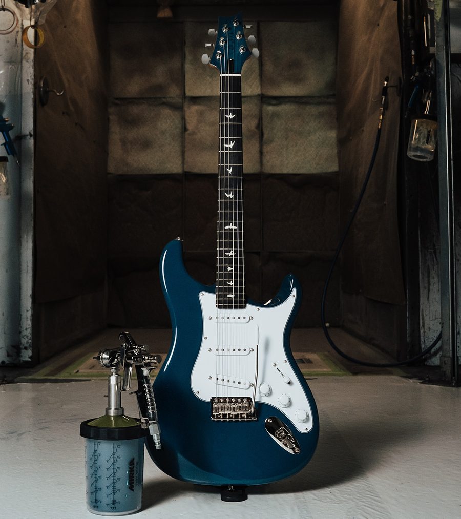 Blue PRS Silver Sky Defender guitar leaning against a garage wall beside a paint spray gun and paint cup